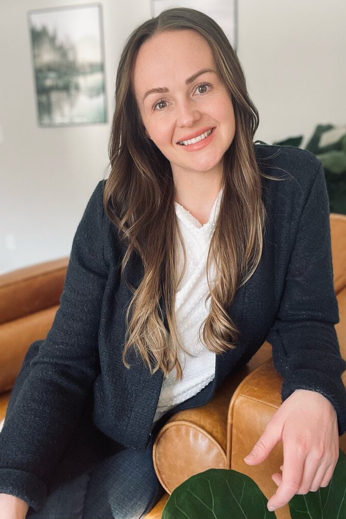 Katie McInnes registered nurse headshot sitting on light brown leather couch in juniper clinic with light walls framed images and plant in background