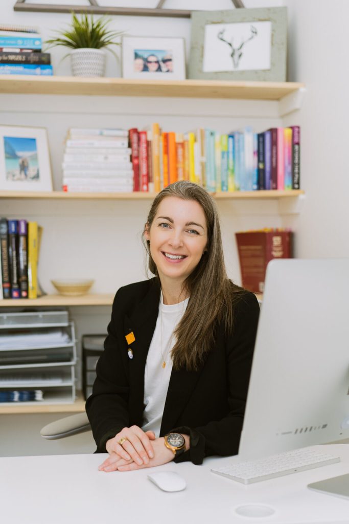 dr. jacinta sullivan naturopathic doctor in fort mcmurray in consult room in front of computer with wall book shelving in the background