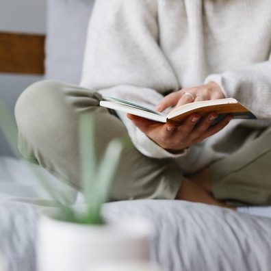 woman in knit sweater reading a book with plant and magazine on table in the forefront