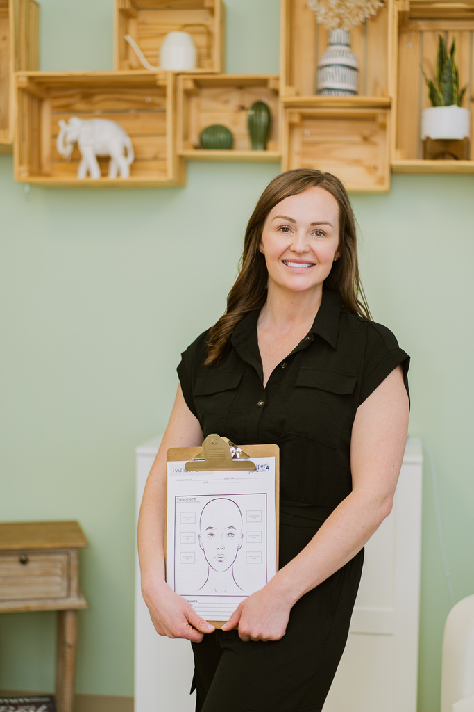 Katie Mcinnes registered nurse aesthetics headshot at juniper naturopathic clinic holding clipboard against green wall background with wooden crate shelving
